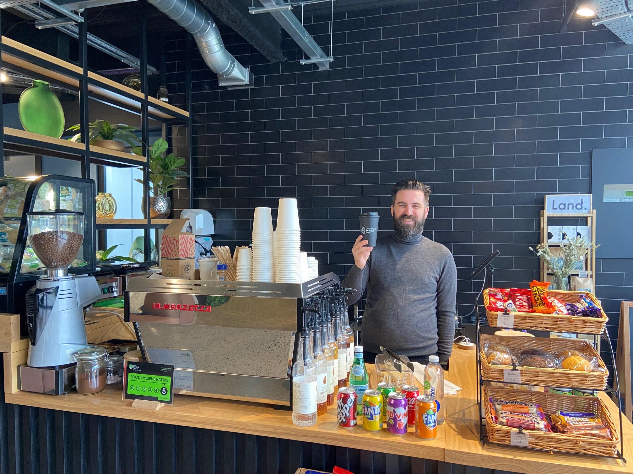 A man stood behind a coffee machine holding a cup of coffee