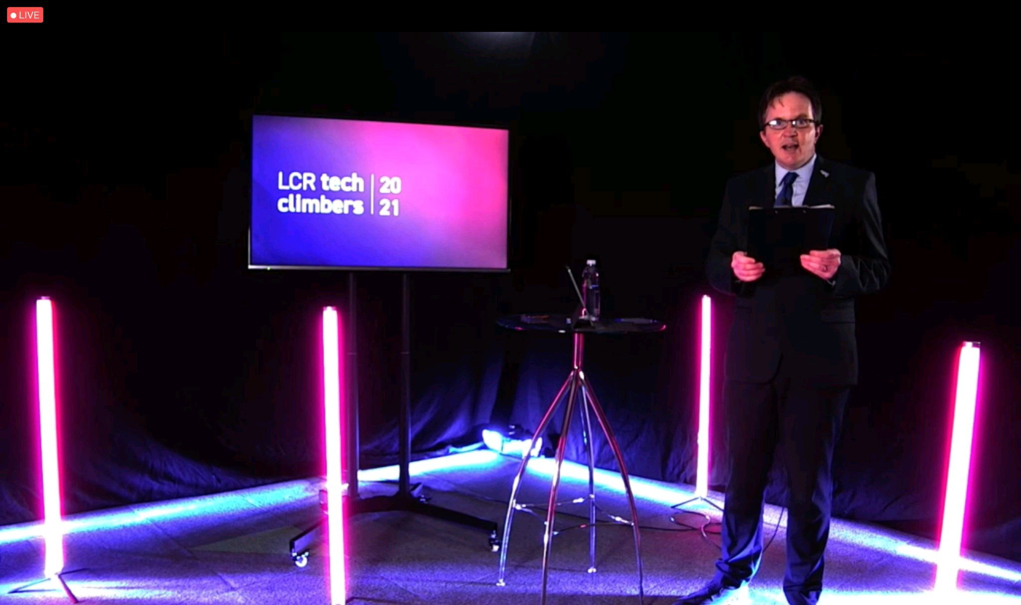 A man giving a talk on a stage with a television next to him
