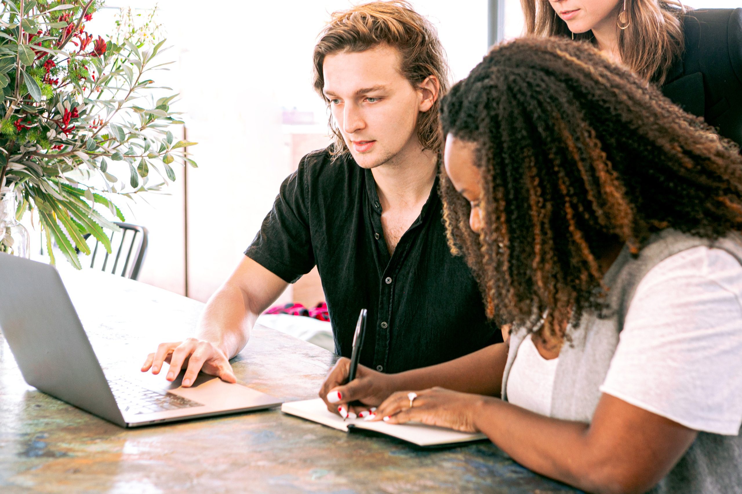 Three people looking at a laptop while one is writing in a notepad